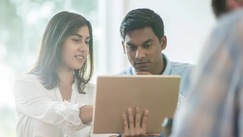 Two colleagues leaning over an open laptop in a bright office, studying the screen together