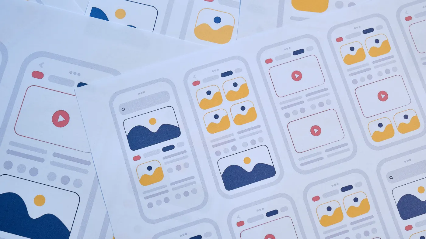 Printed sheets of mobile app wireframes in blue, yellow and red, fanned across a desk