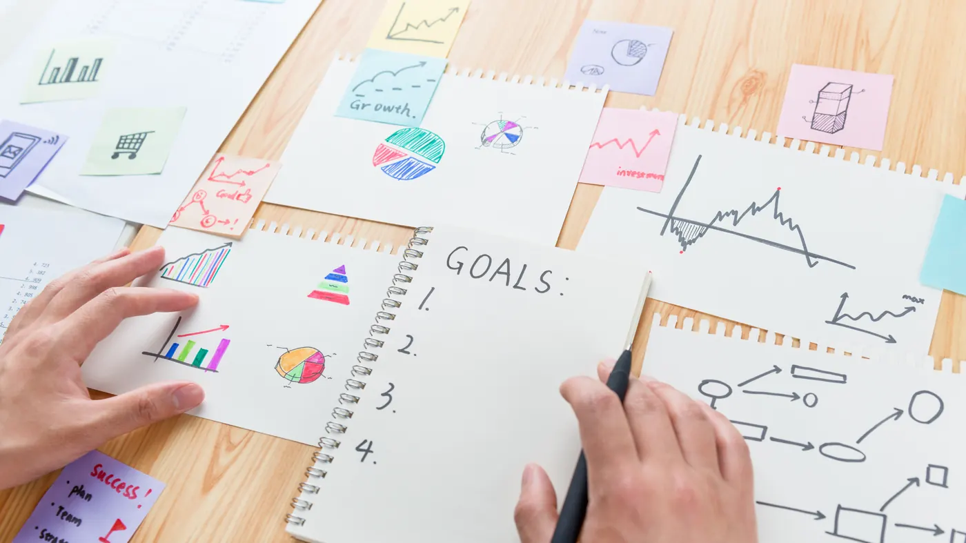 A desk covered with hand-drawn charts and sticky notes, a hand writing a numbered list headed GOALS