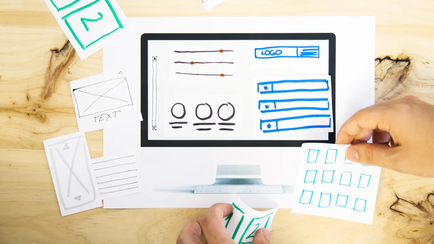 Hand-drawn wireframe cards laid out around a monitor sketch on a wooden desk