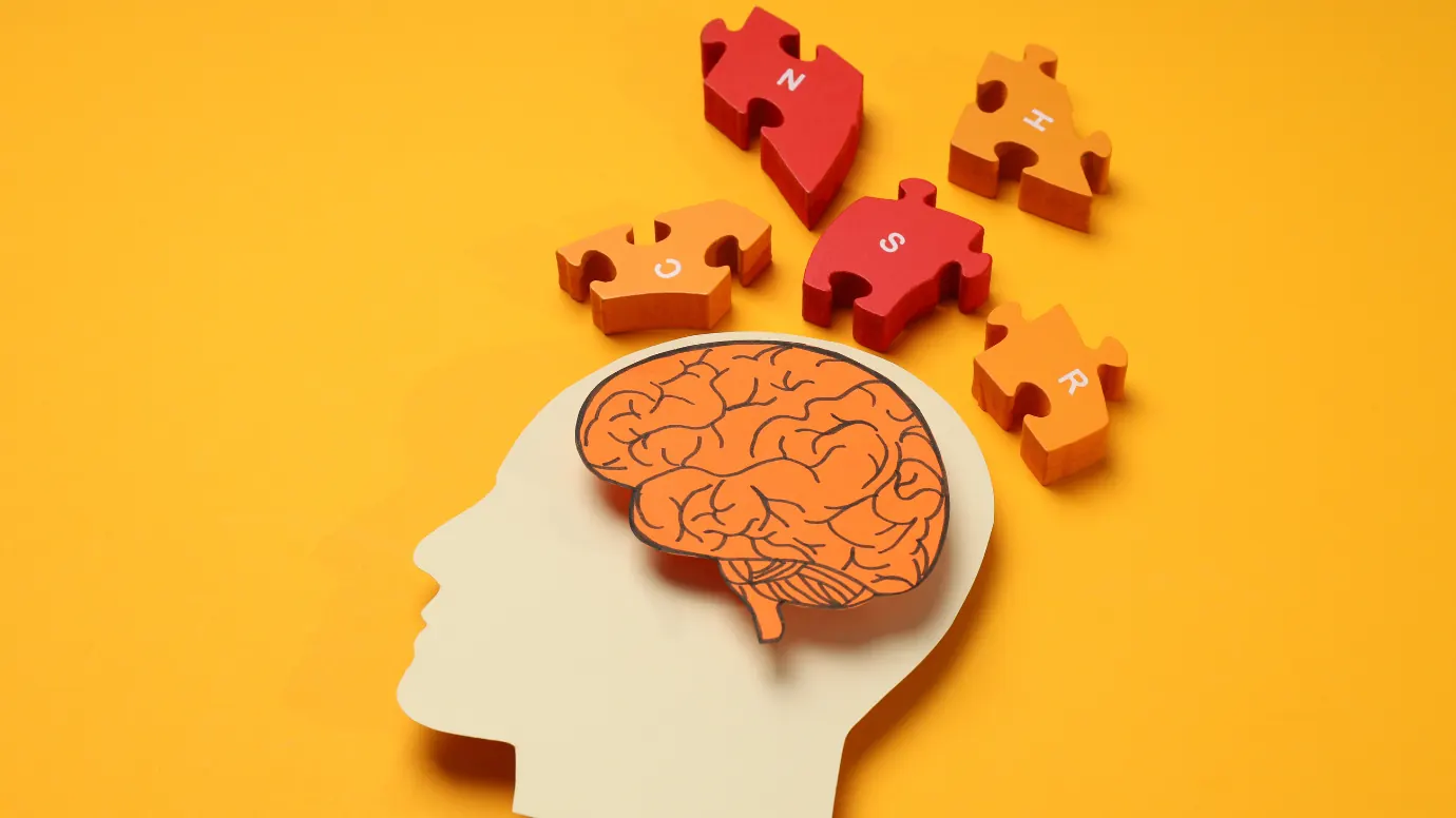 A paper cut-out head in profile with an orange brain, jigsaw pieces scattering above it