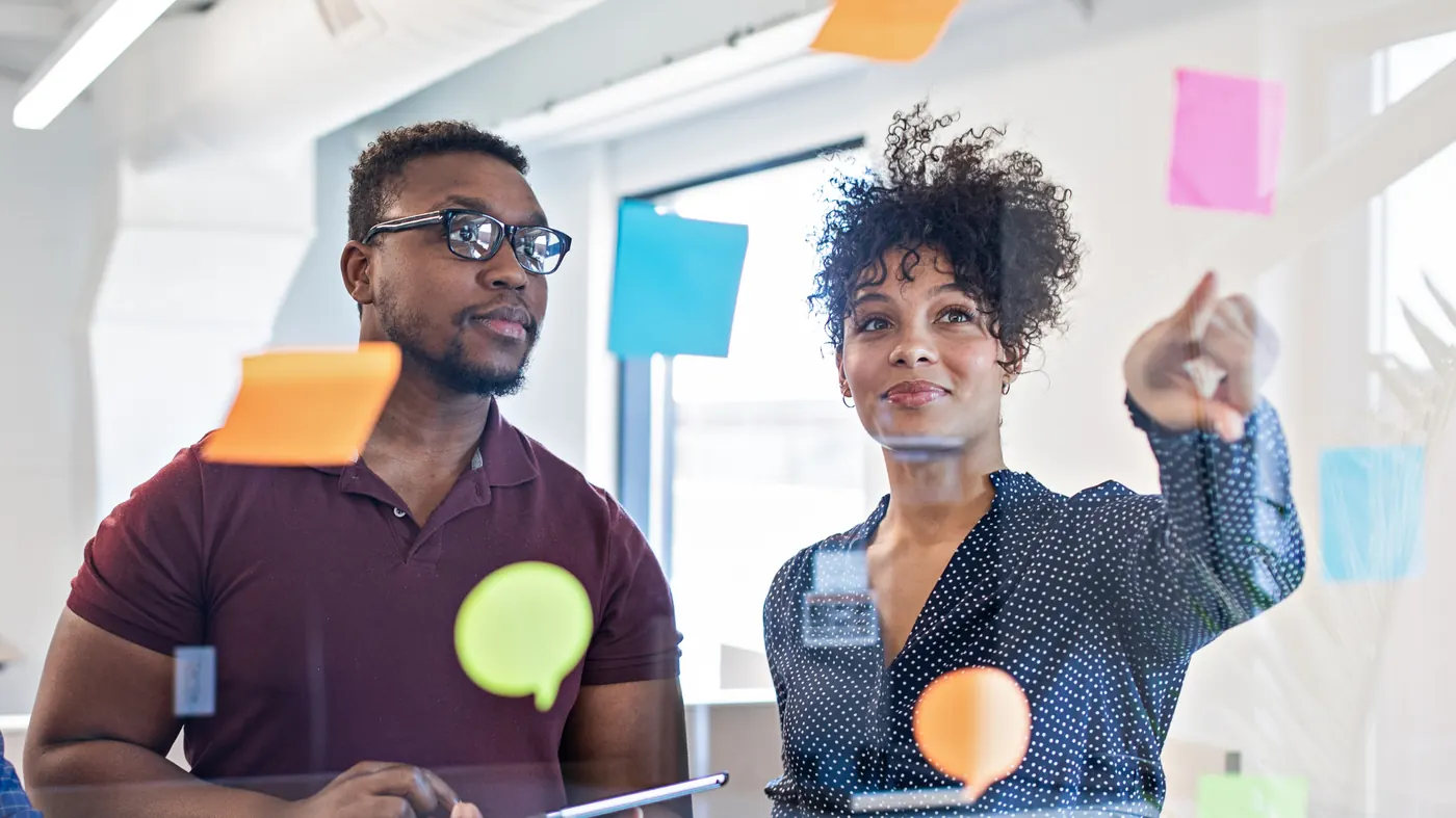 Two colleagues adding sticky notes to a glass wall in an office, one of them pointing at the board