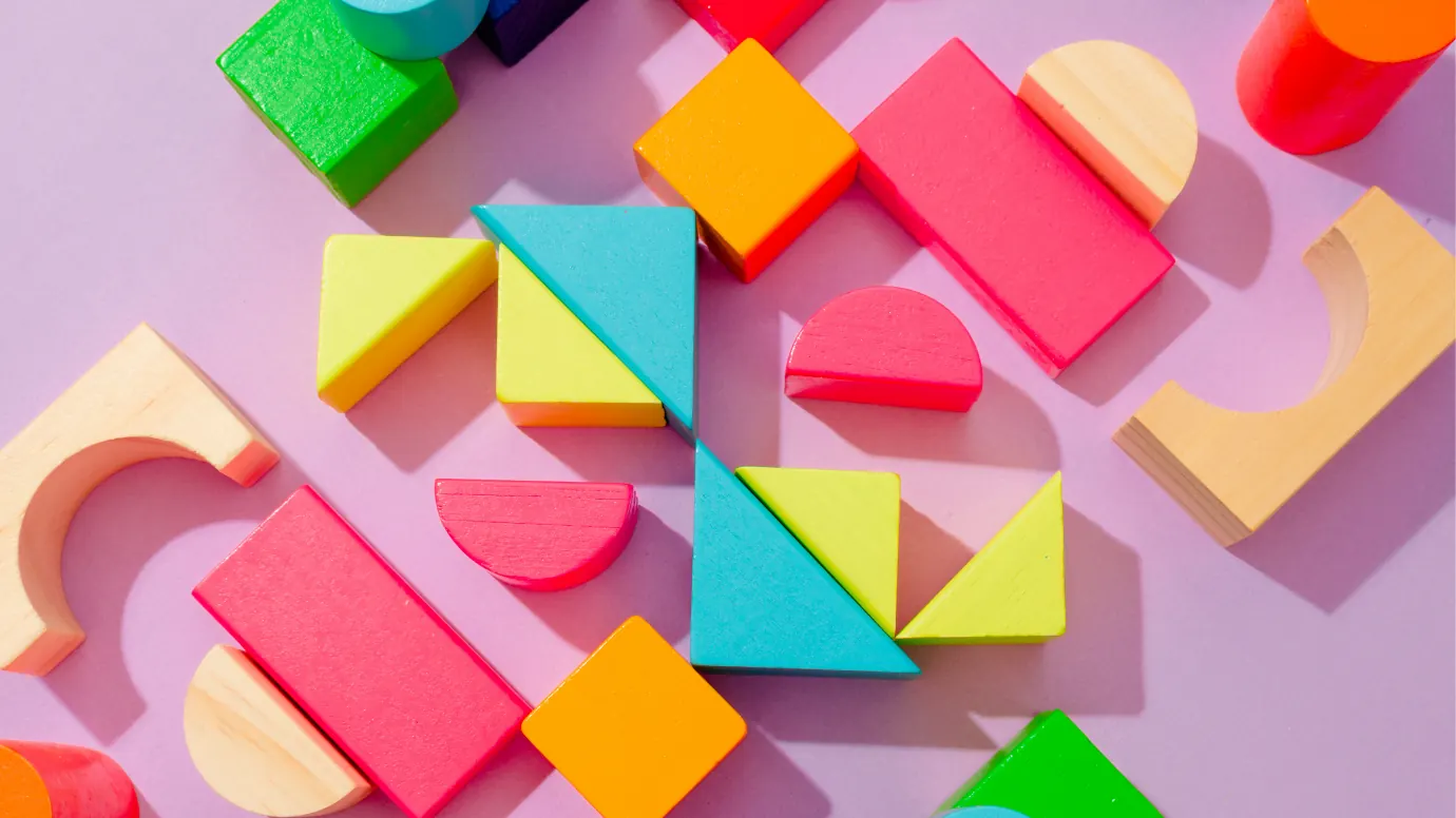Coloured wooden blocks — cubes, arcs, triangles and half-discs — scattered across a pink surface, with several fitted together into one larger square