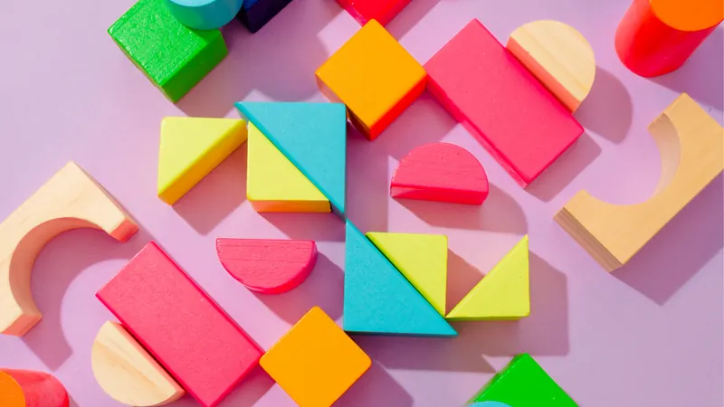 Coloured wooden blocks — cubes, arcs, triangles and half-discs — scattered across a pink surface, with several fitted together into one larger square