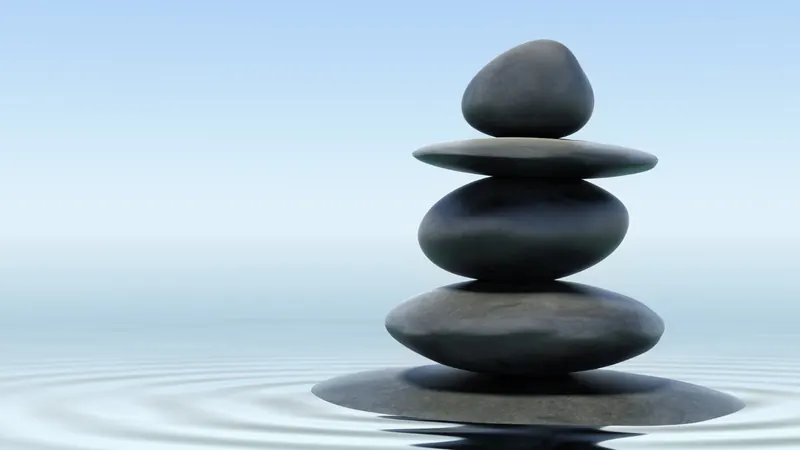 A cairn of five smooth dark stones balanced above still water, with ripples spreading out around it